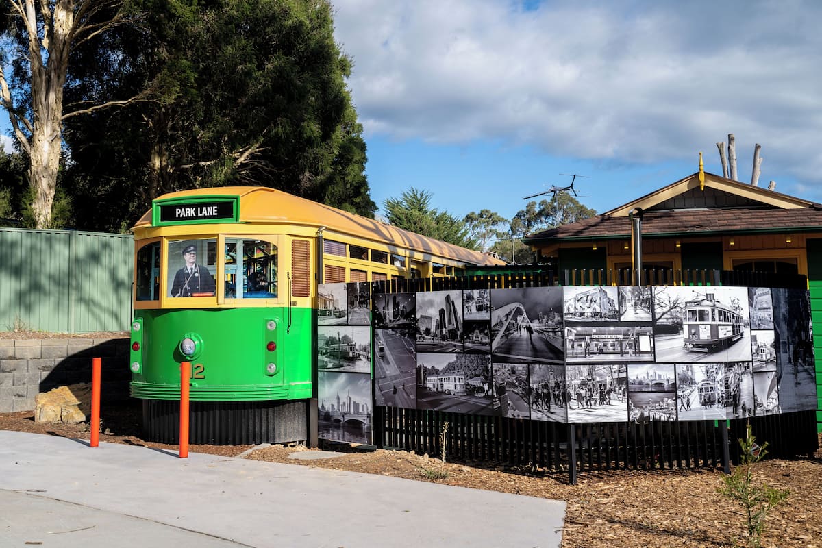 A tram with a green and yellow exterior, parked next to a fence with historical photos.