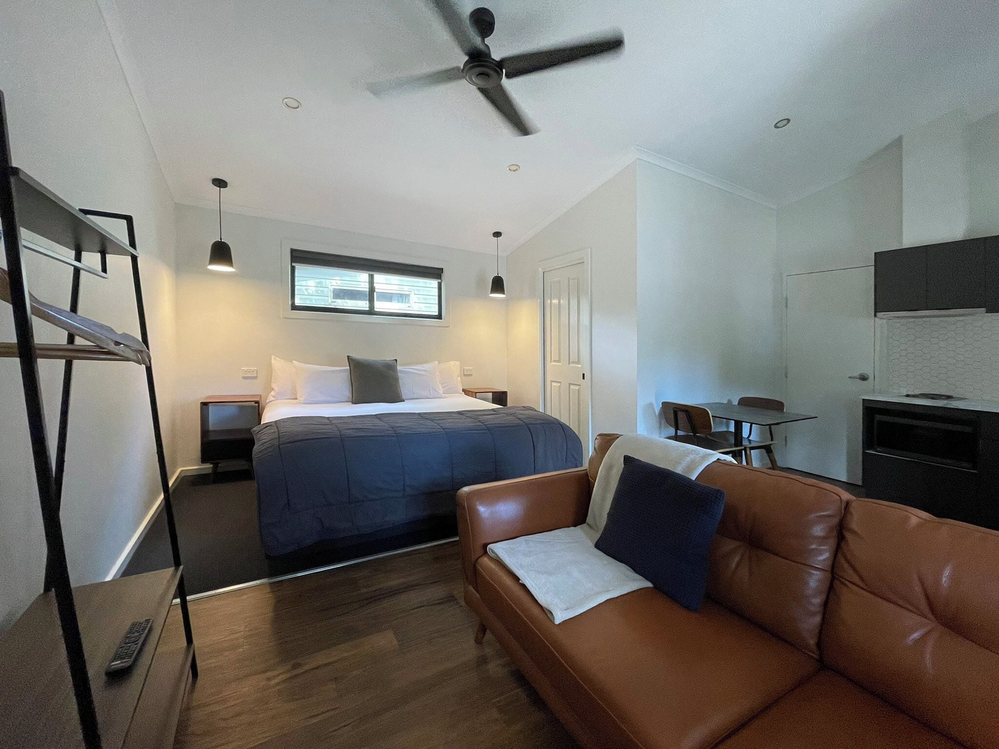 Une chambre d’hôtel compacte comprenant un lit, un canapé en cuir marron, une petite table, un coin cuisine et un ventilateur de plafond.