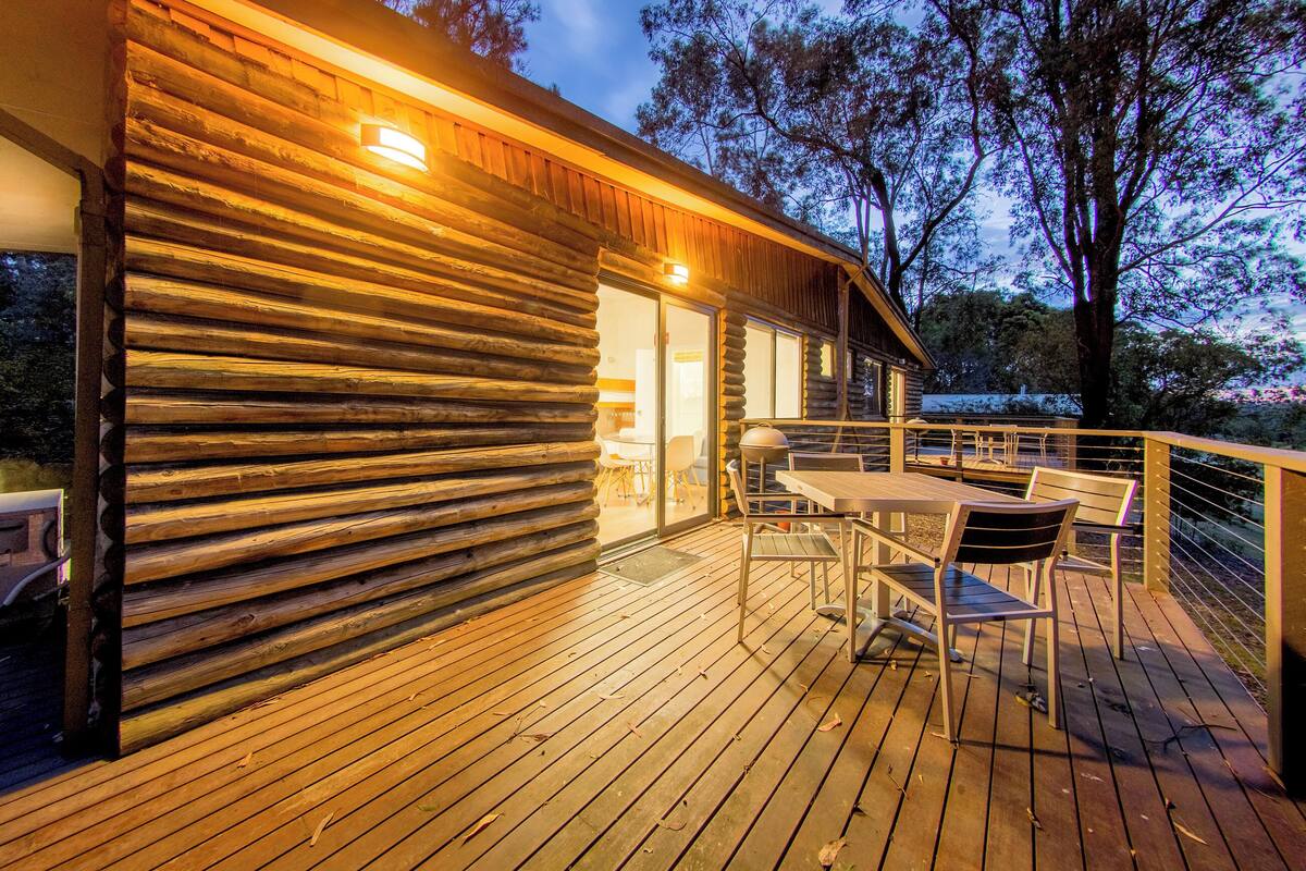 A wooden cabin with a deck, outdoor seating, and a view of trees.