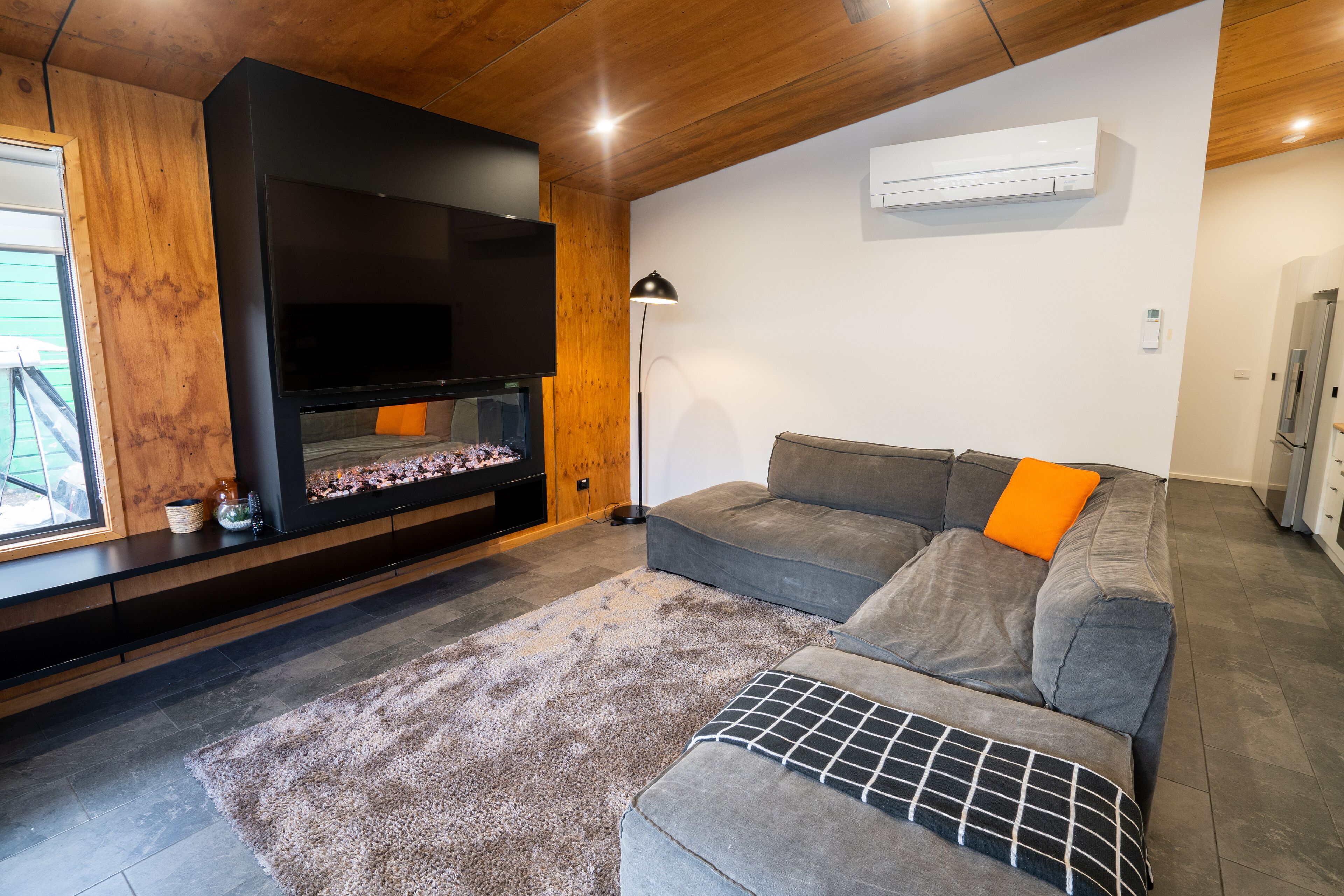 A modern living room with a flat-screen TV, a gray sofa with an orange cushion, a patterned rug, and wooden ceiling beams.