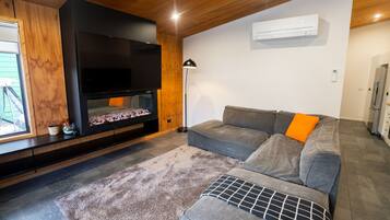 A modern living room with a flat-screen TV, a gray sofa with an orange cushion, a patterned rug, and wooden ceiling beams.