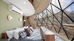 Un dormitorio con cama, almohadas, una mesita de noche, una lámpara y vistas a través de un ventanal grande.