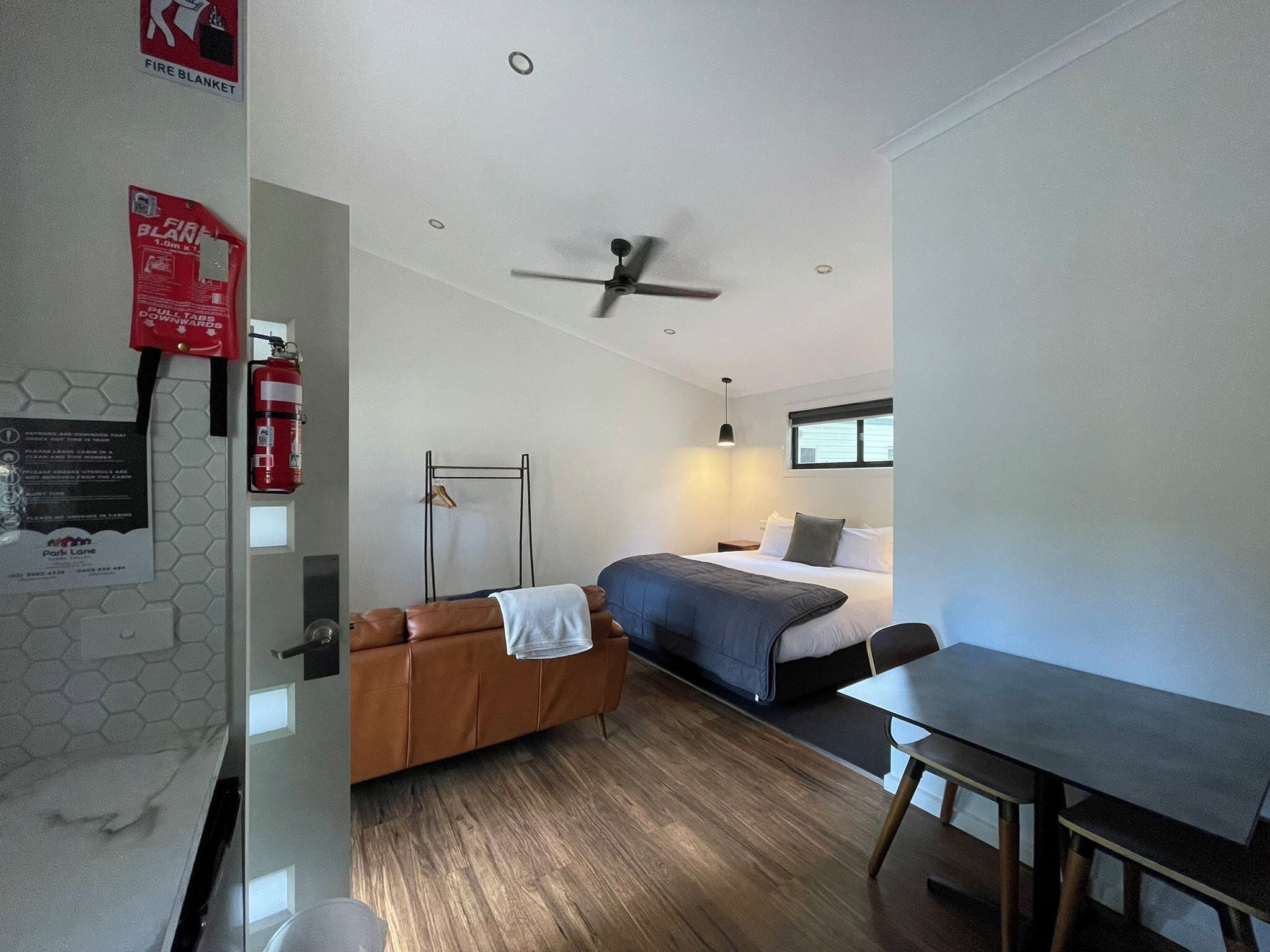 A modern hotel room with a bed, a desk, a chair, a ceiling fan, and a fire extinguisher.