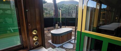 Une porte en bois donne accès à un patio équipé d’un jacuzzi, bordé d’un chemin de pierres et offrant une vue sur une colline boisée.