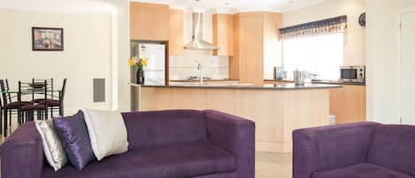A modern living room with a purple sofa, a glass coffee table, and a dining area with chairs.