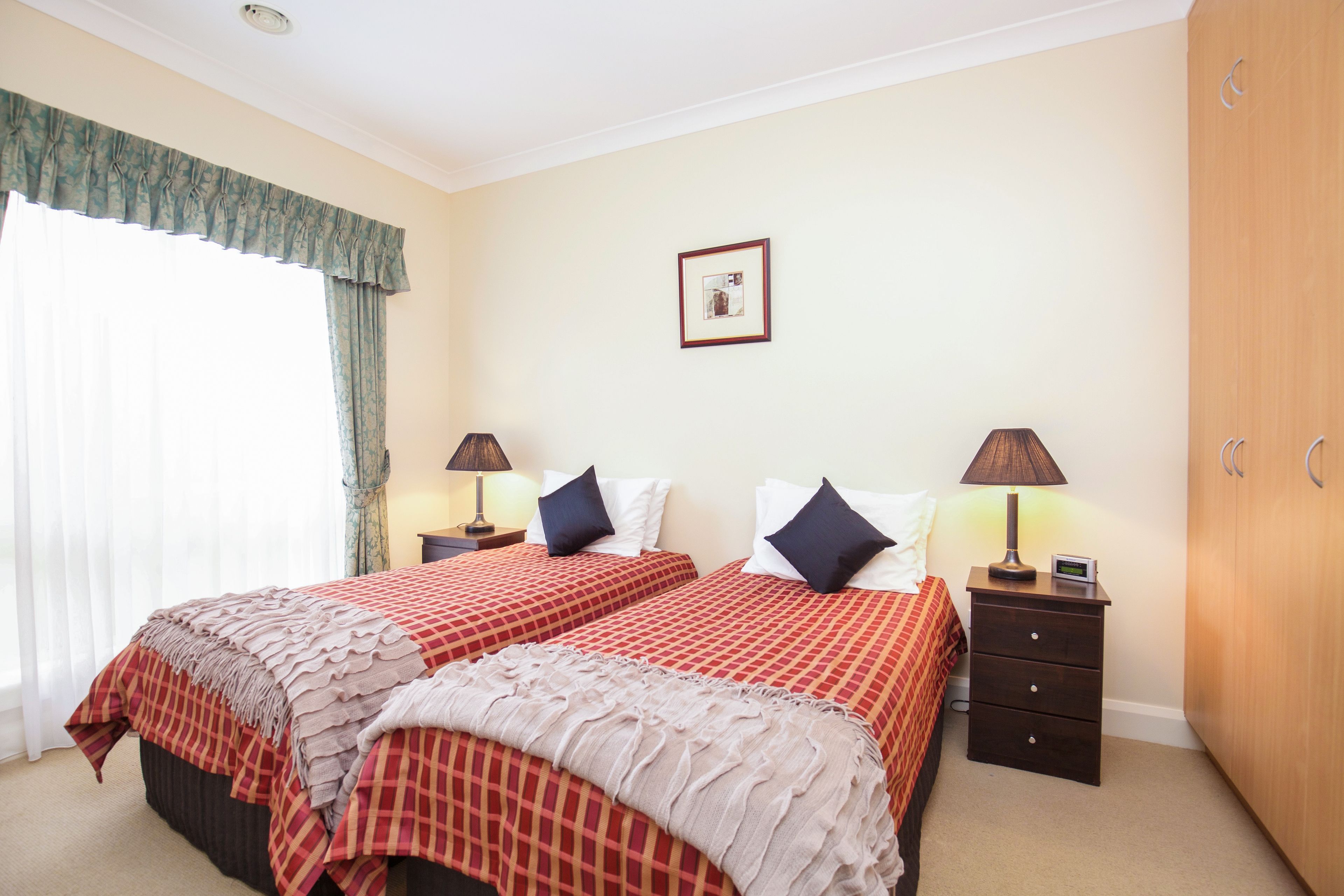 Een hotelkamer met twee bedden, elk voorzien van een rood-wit geruit sprei, twee nachtkastjes met lampen, een houten kledingkast en een ingelijst schilderij aan de muur.