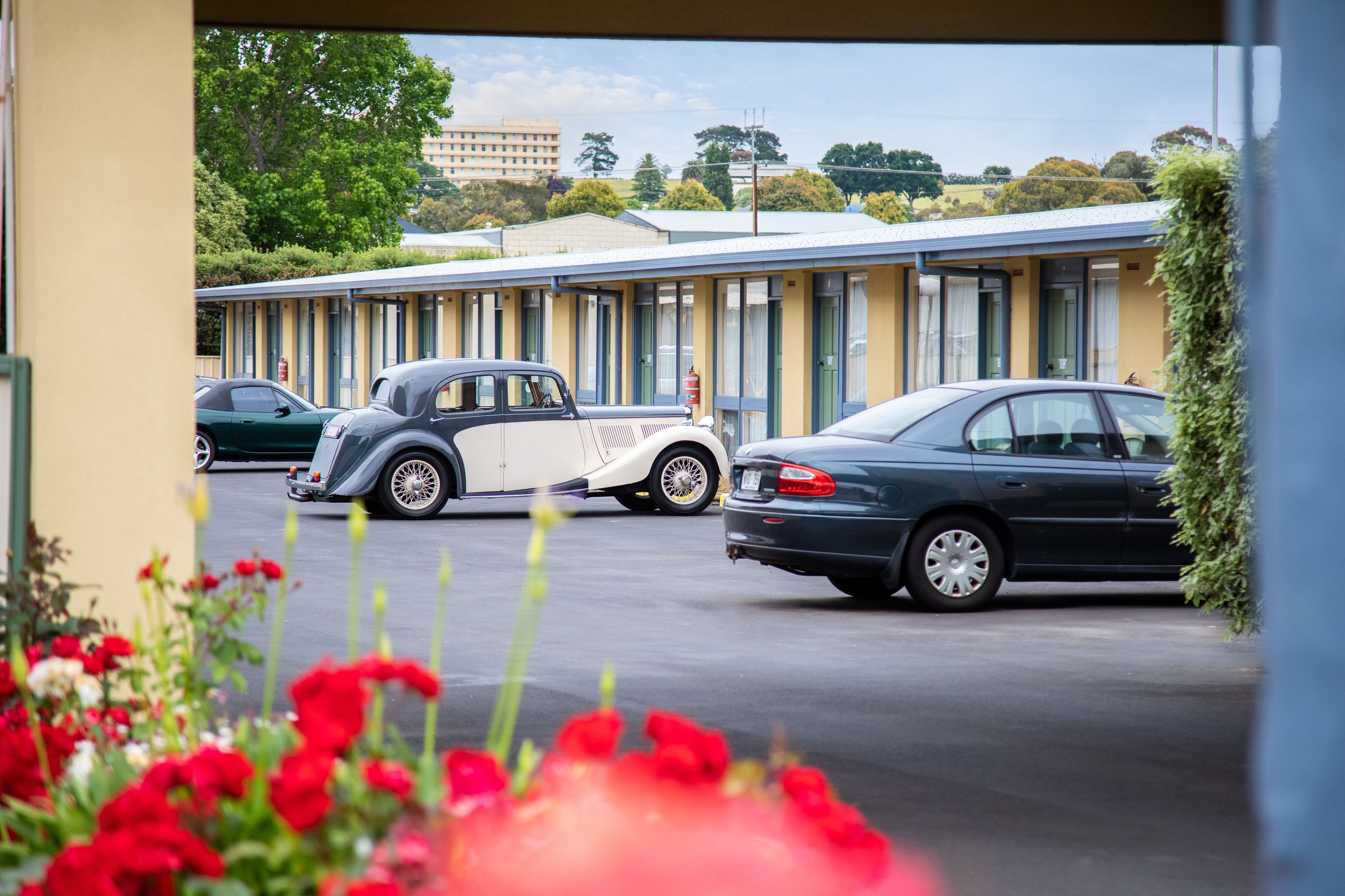 Um motel com uma fileira de quartos, um estacionamento com vários carros e uma construção ao fundo.