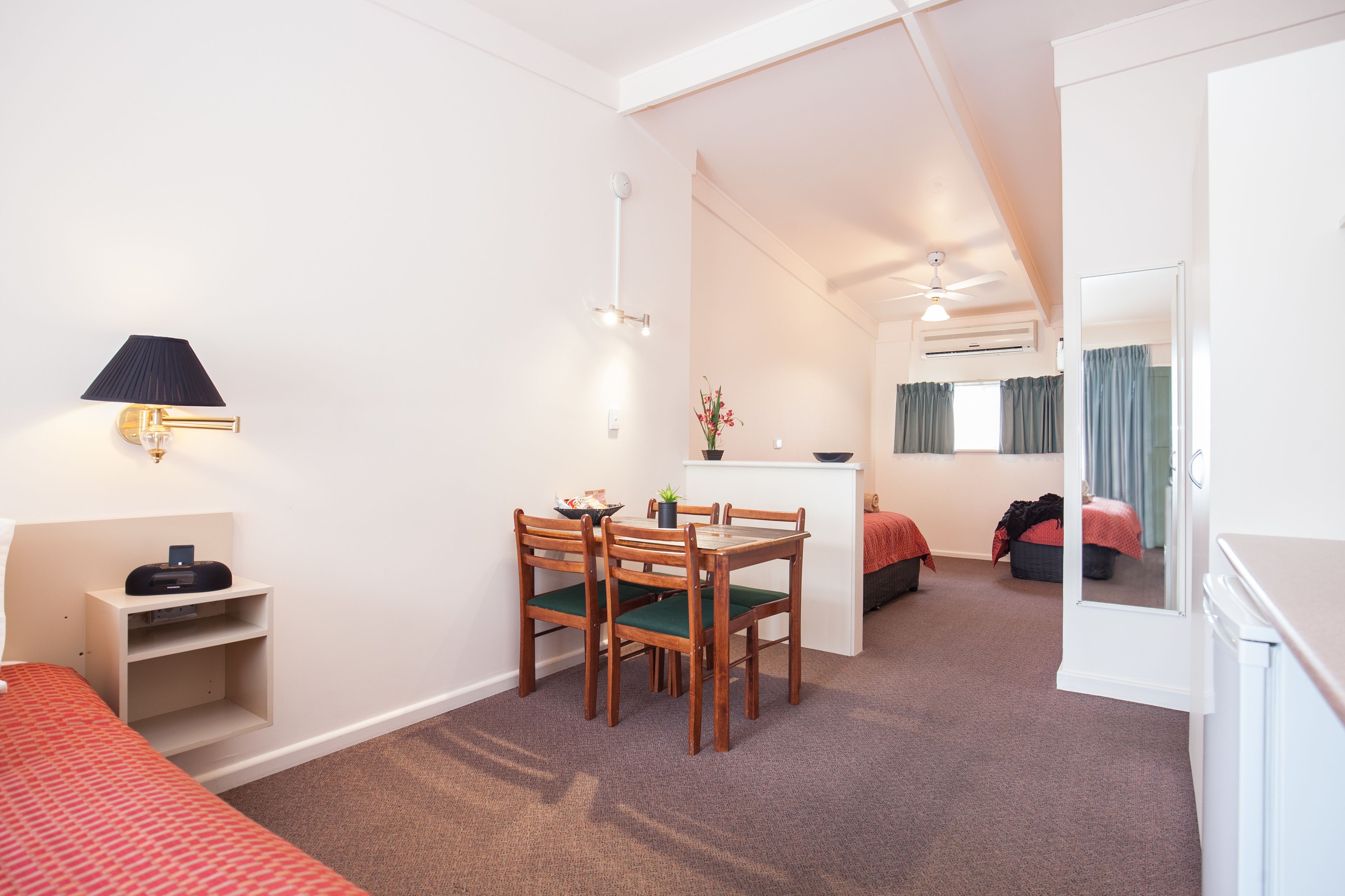 A room with a bed, a desk with a lamp, a chair, a small table with a plant, and a mirror.