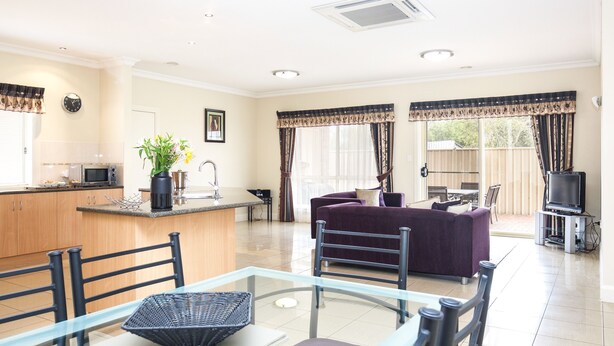 A modern living room with a glass dining table, purple sofa, and a kitchen area with a microwave. - Mid City Motel - Mount Gambier (Mount Gambier)
