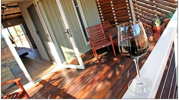 An outdoor patio with wooden flooring, a glass table with a wine glass, a red chair, and a wooden fence. - Mungo Lodge (Arumpo)