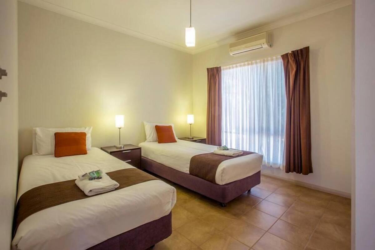 A hotel room with two beds, a nightstand with a lamp, a window with curtains, and an air conditioning unit.