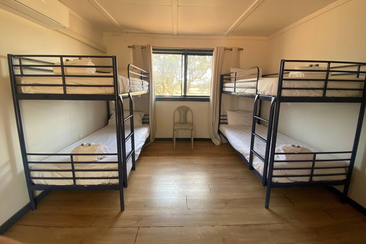 A room with four bunk beds arranged in a row, a window with curtains, a chair, and a wooden floor.