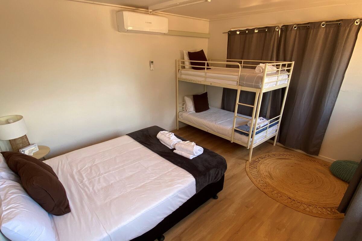 A room with a bunk bed, a single bed, a bedside table with a lamp, and a round woven rug.
