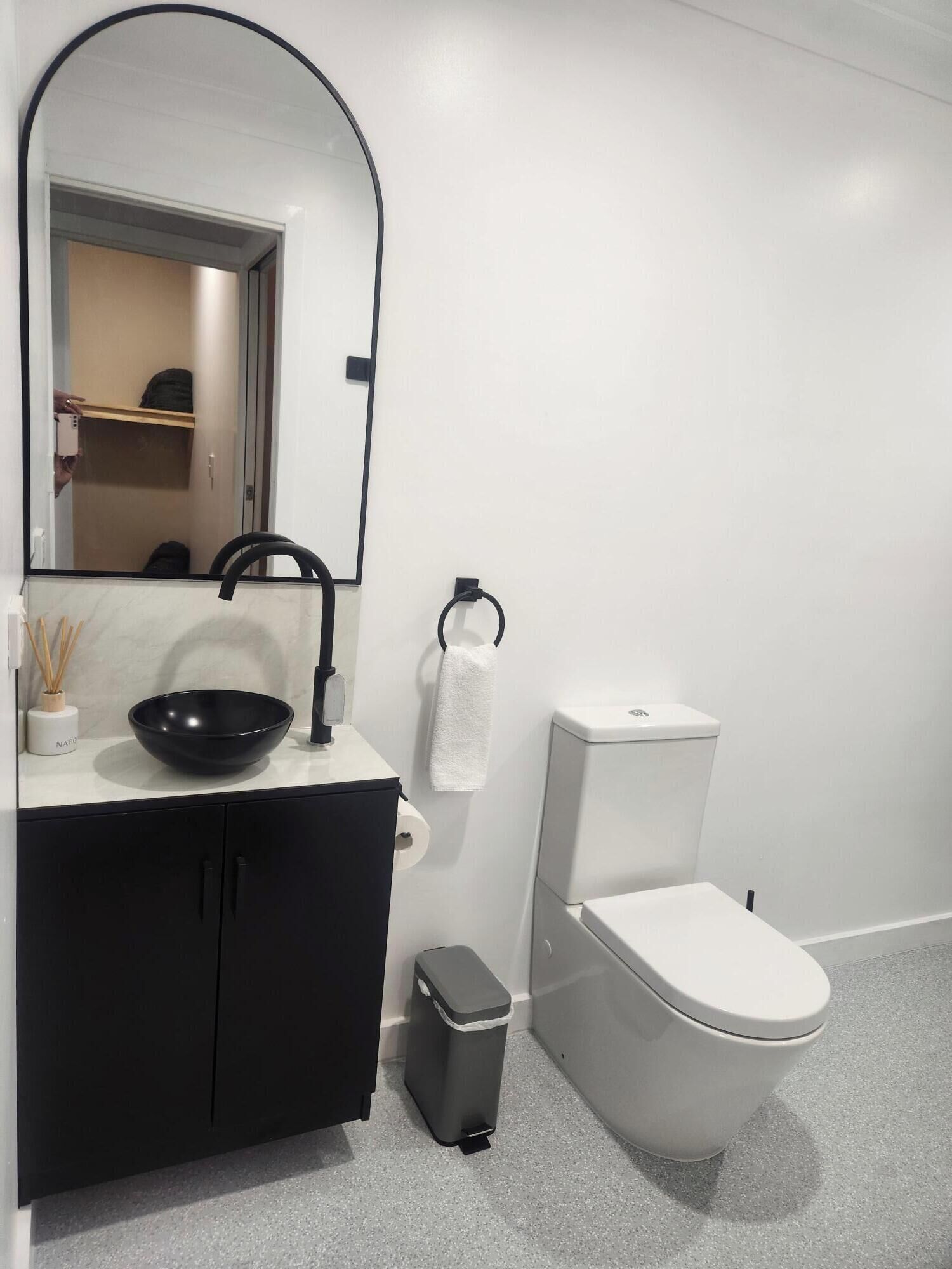 A modern bathroom with a black vanity, a large mirror, a black bowl on the counter, a white toilet, and a trash bin.