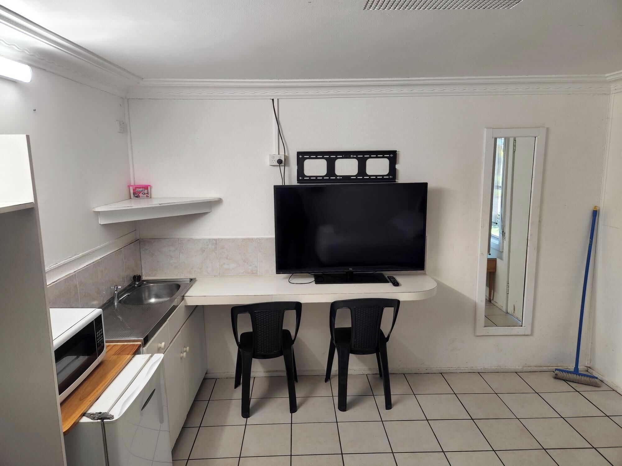 A compact kitchen with a microwave, sink, and two chairs. A flat-screen TV is mounted above the counter.