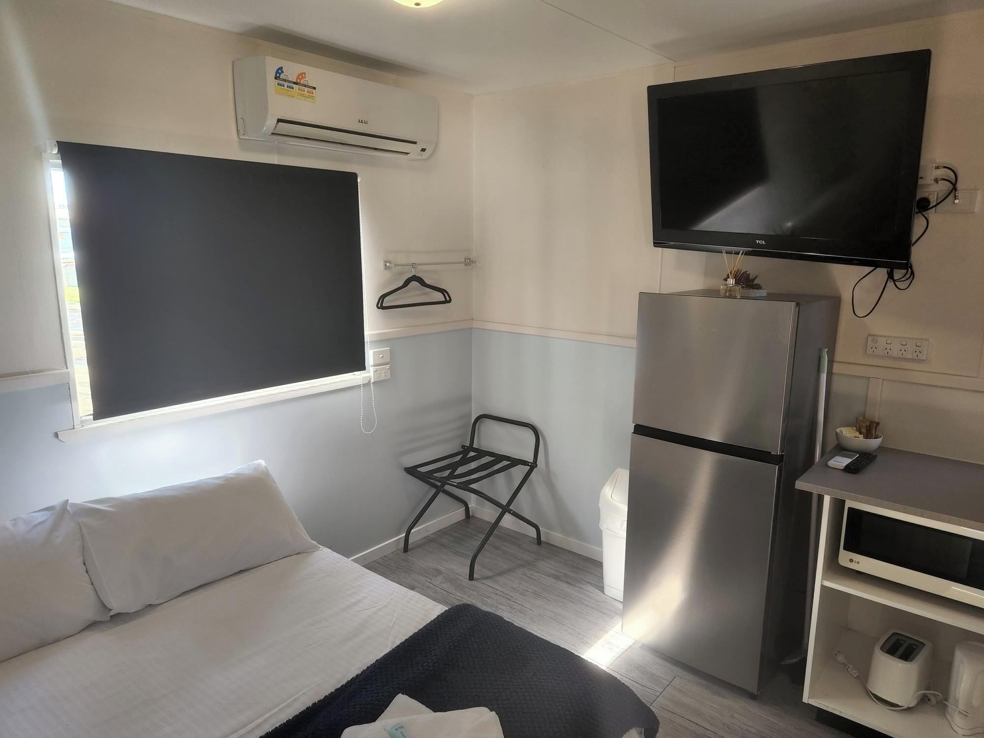 Une chambre d’hôtel compacte comprenant un lit, une petite kitchenette, un téléviseur fixé au mur et une fenêtre avec un store occultant noir.