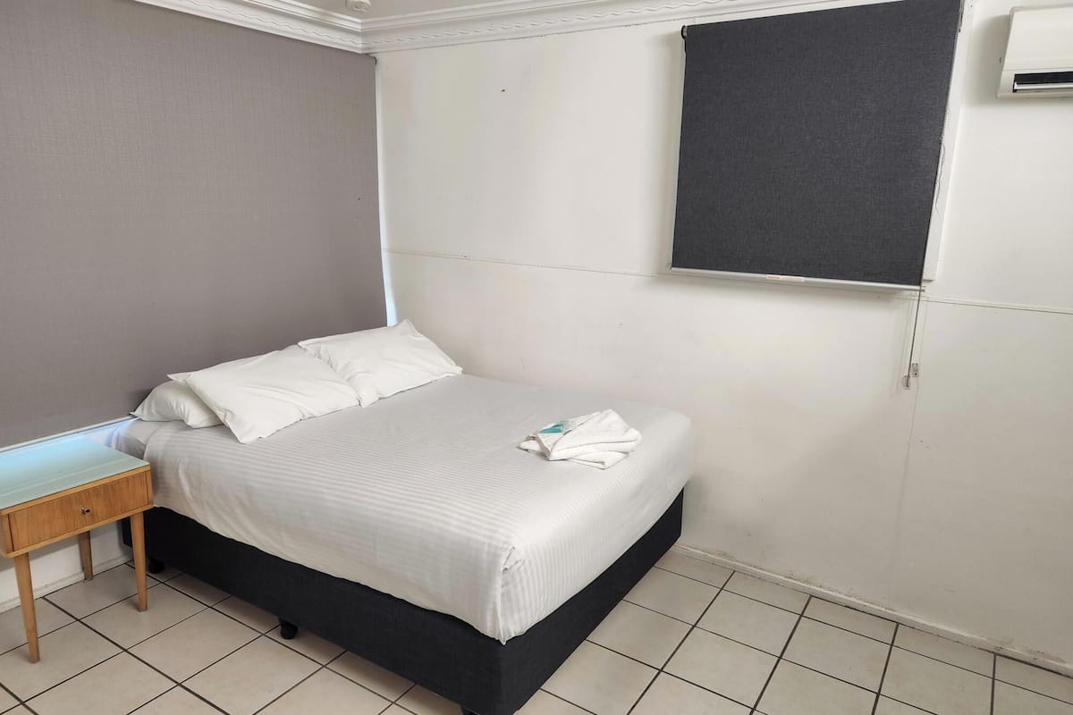 A small, clean room with a single bed, a bedside table, and a window with a dark blind.