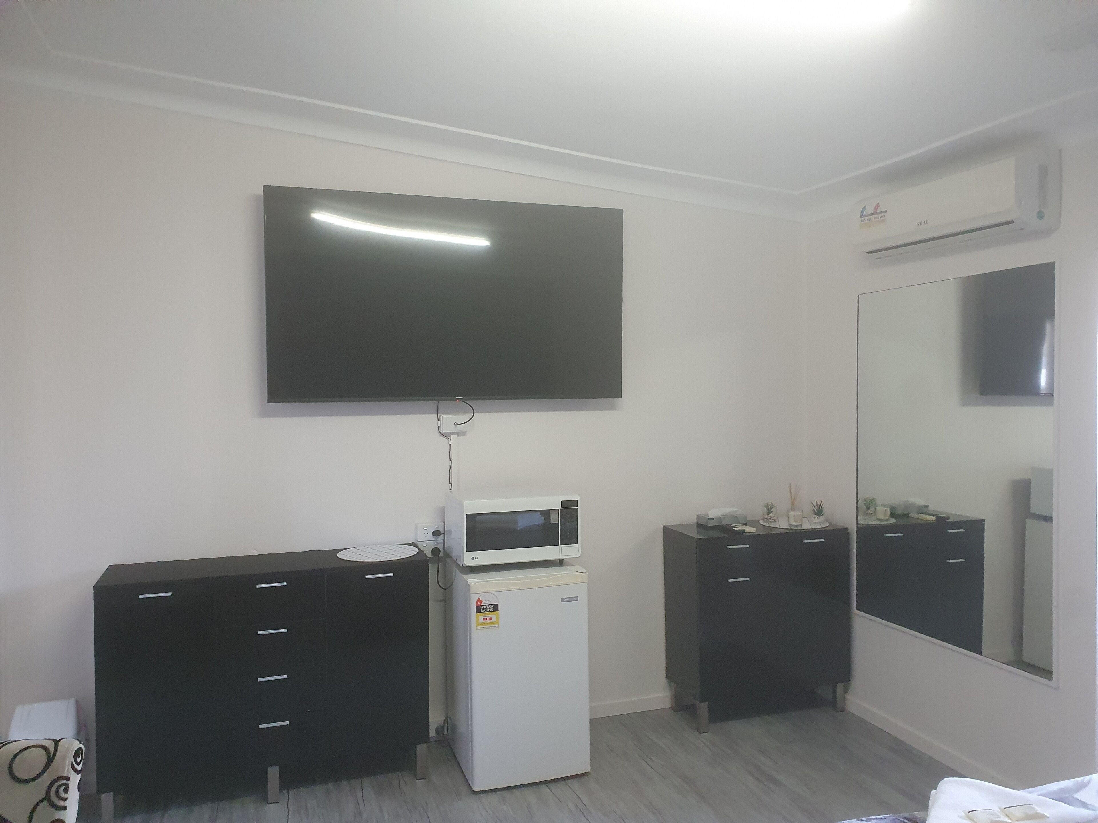 A room with a flat-screen TV mounted on the wall, a small refrigerator, a microwave, a black dresser, and a mirror.