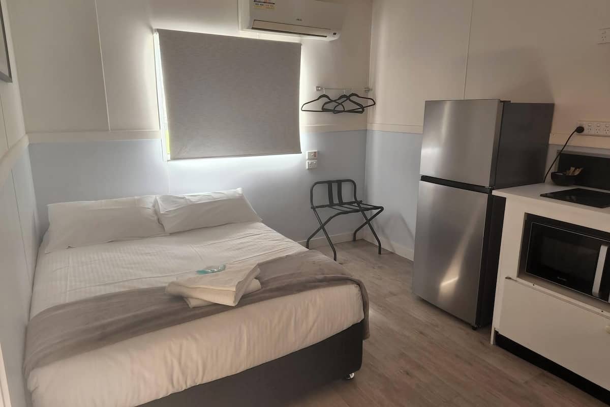 A compact hotel room with a bed, a small kitchenette, a window with blinds, and a wall-mounted air conditioner.