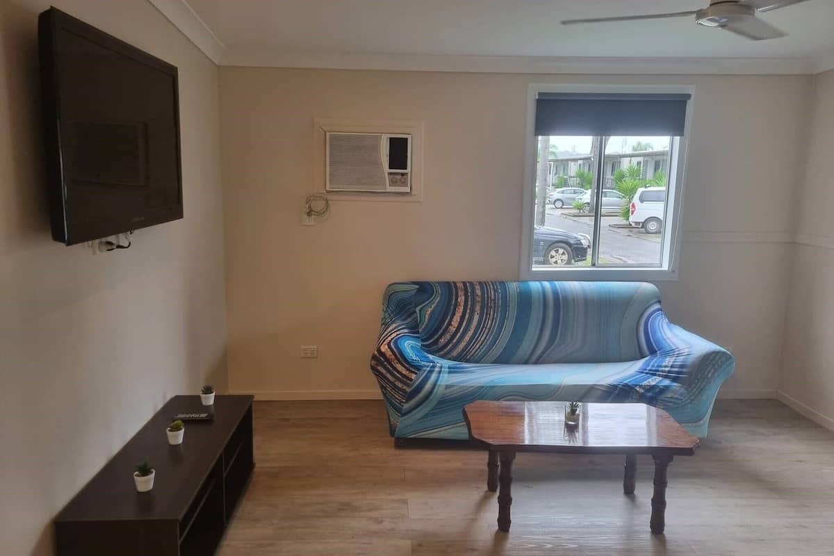 A living room with a sofa, a coffee table, a TV mounted on the wall, a window with a view of the street, and a ceiling fan.