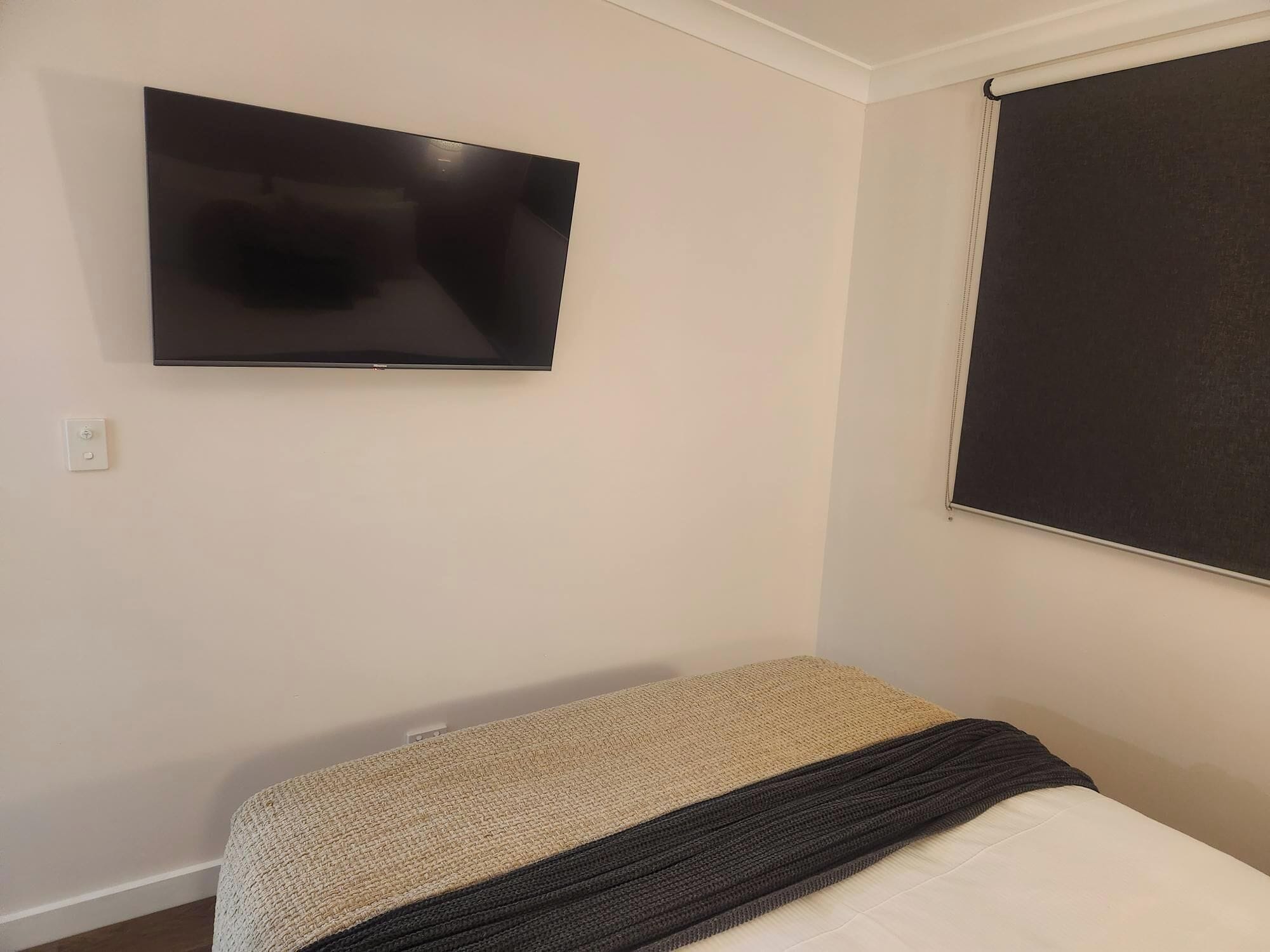 A bedroom with a bed, a flat-screen TV mounted on the wall, and a window with a blind.