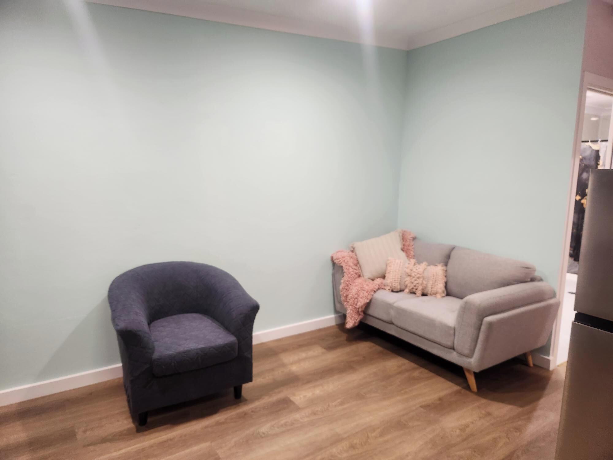 A room with a dark armchair, a grey sofa with pink throw pillows, and a wooden floor.