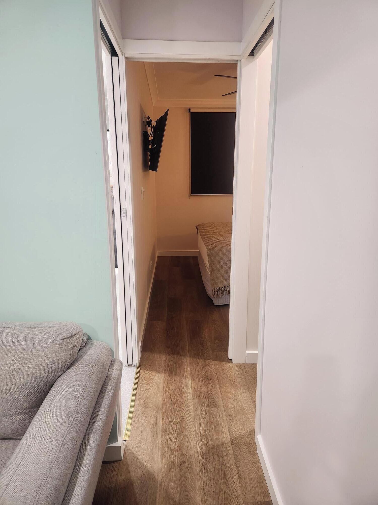 A narrow hallway with a bed at the end, a wall-mounted TV, and a door leading to another room.