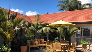 Un patio con un banco y una mesa de madera, rodeado de macetas con plantas y un paraguas amarillo.