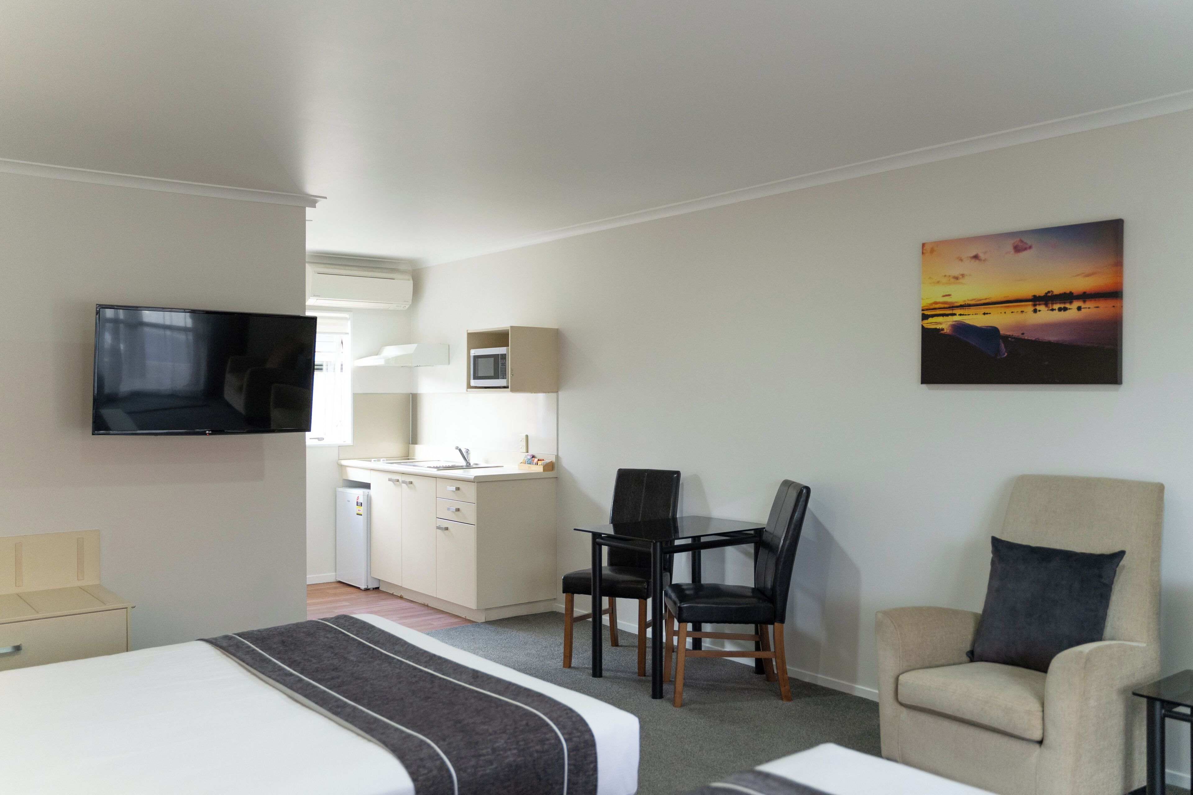 Habitación de hotel con cama, escritorio con dos sillas, televisor, microondas y un cuadro de un atardecer.