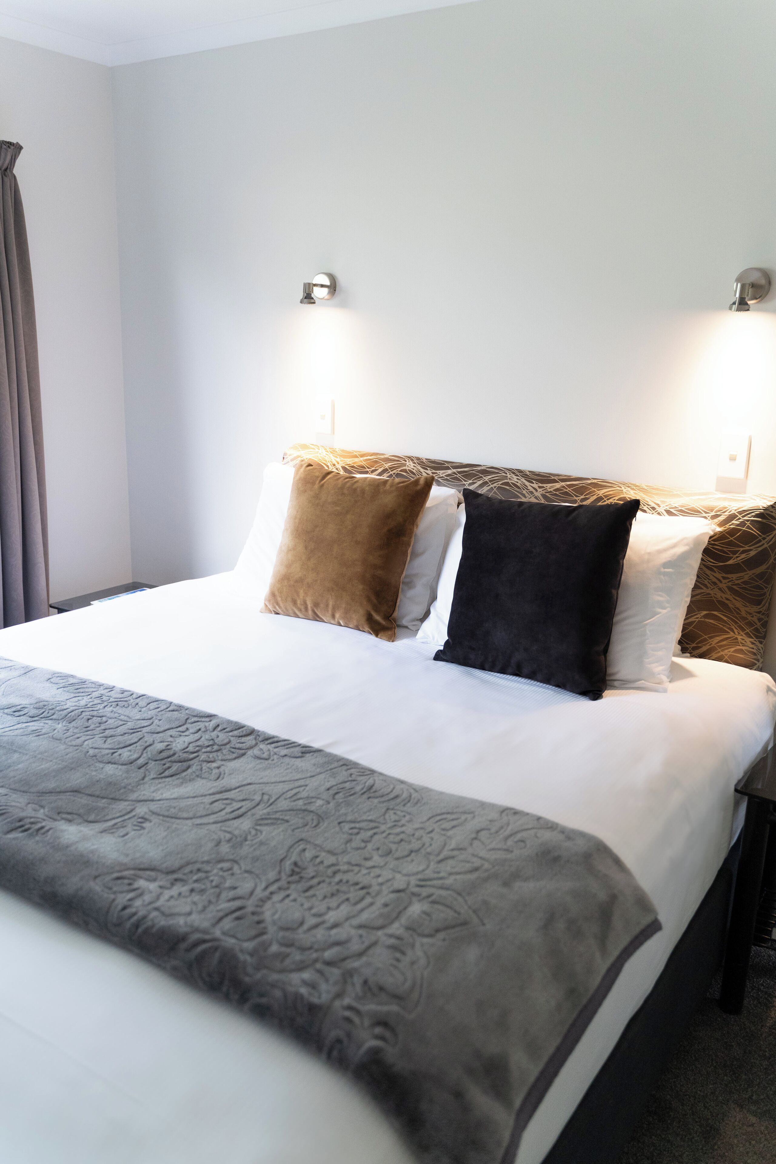 A neatly made bed with a patterned throw, two wall-mounted reading lights, and a window with curtains.