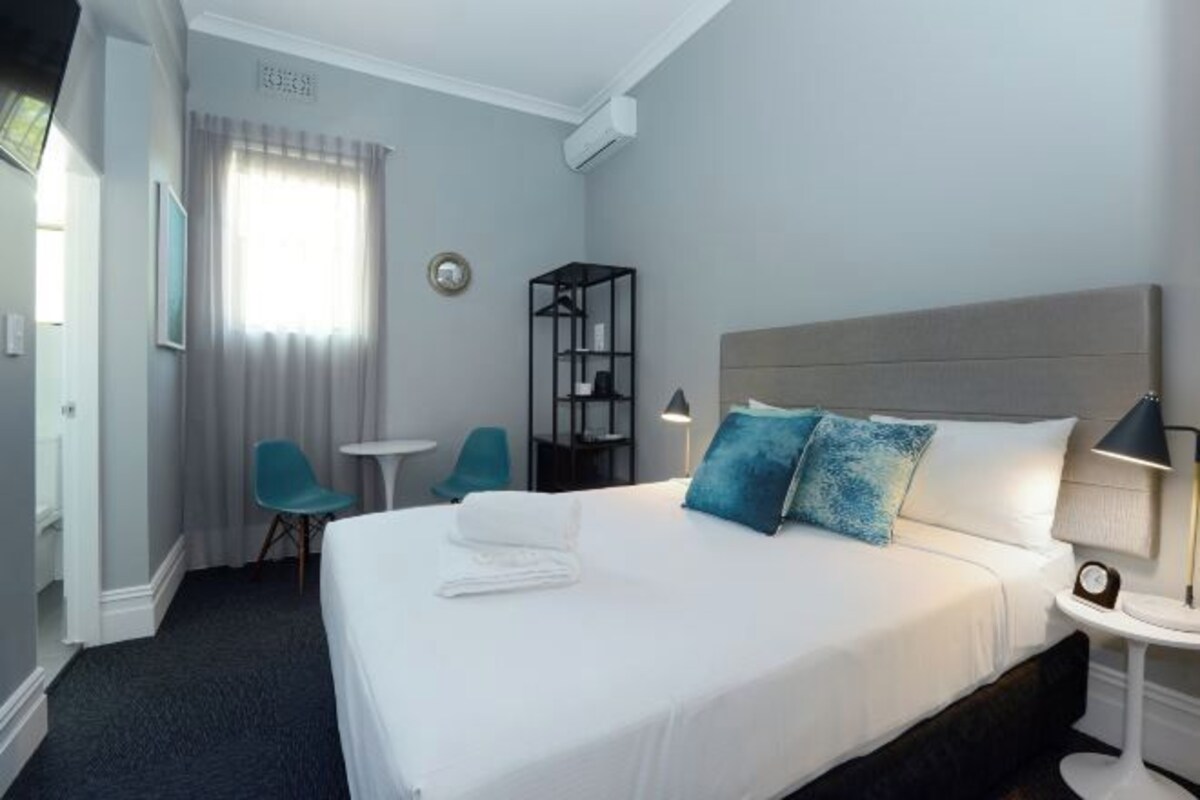 Ein Hotelzimmer mit einem großen Bett, einem kleinen Tisch, einem Stuhl und einem an der Wand befestigten Regal.