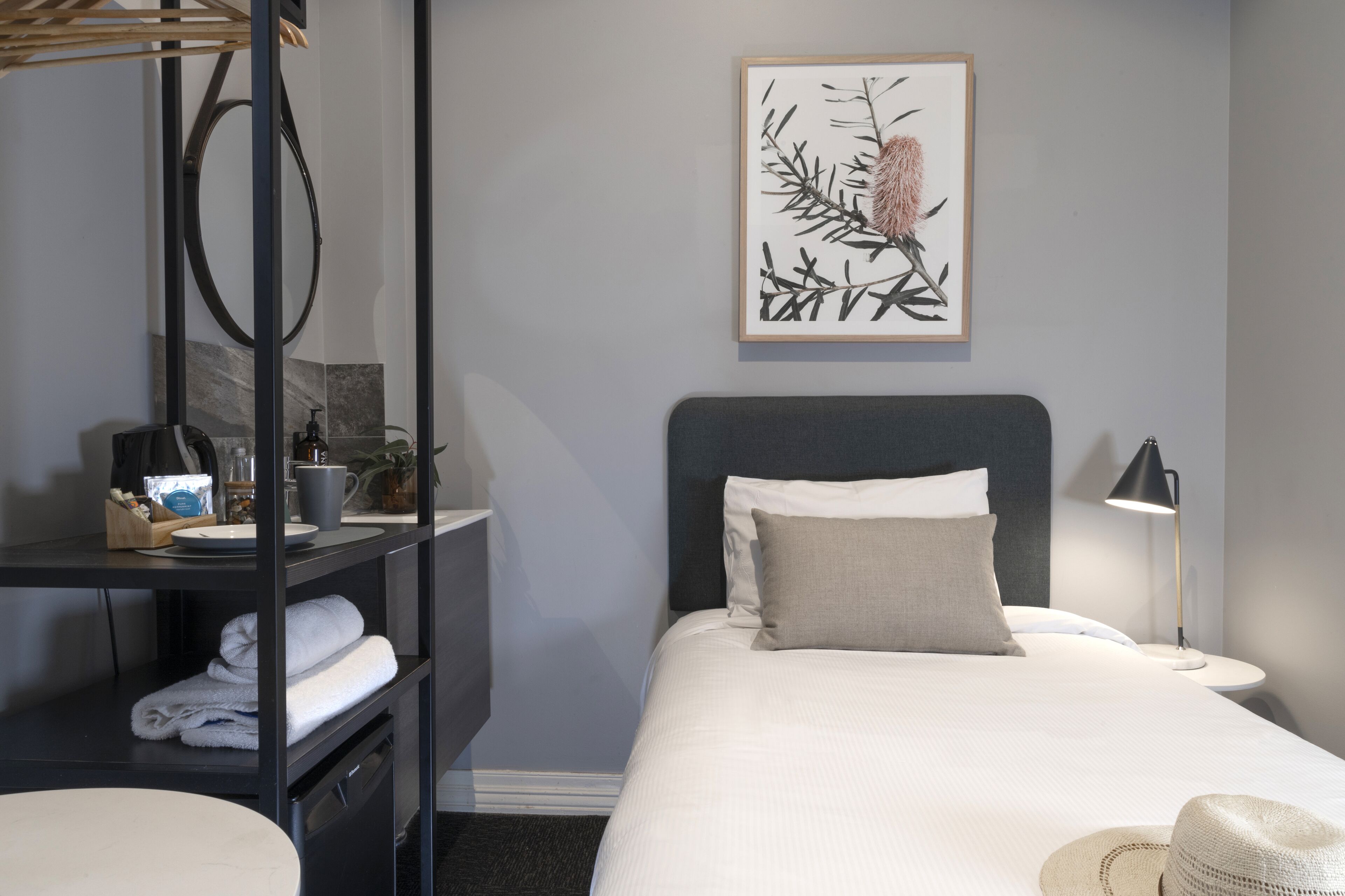 A hotel room with a bed, a nightstand, a desk with a lamp, a shelf with towels, and a framed artwork on the wall.