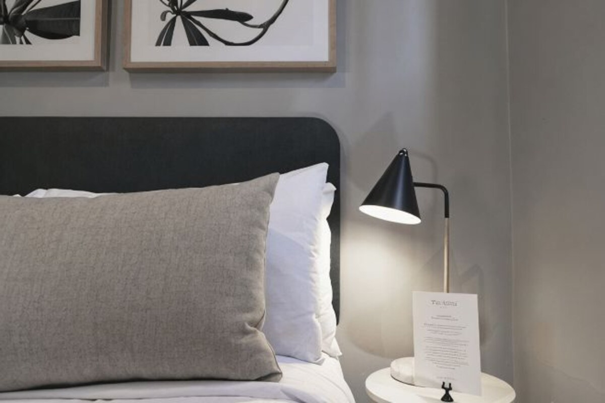 Ein Bett mit schwarzem Kopfteil, einem grauen Kopfkissen, einem weißen Kopfkissen, einer schwarzen Schreibtischlampe und einem kleinen Tisch mit einem Dokument.