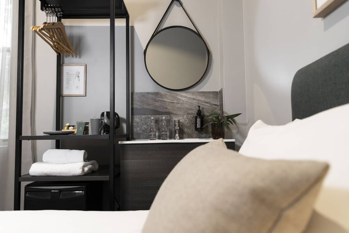 A modern hotel room with a bed, a bedside table with a lamp, a mirror, and a shelf with toiletries.