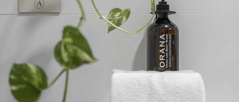Un baño con un lavabo blanco, una botella de ORANA para lavado de manos y cuerpo, y una toalla blanca.