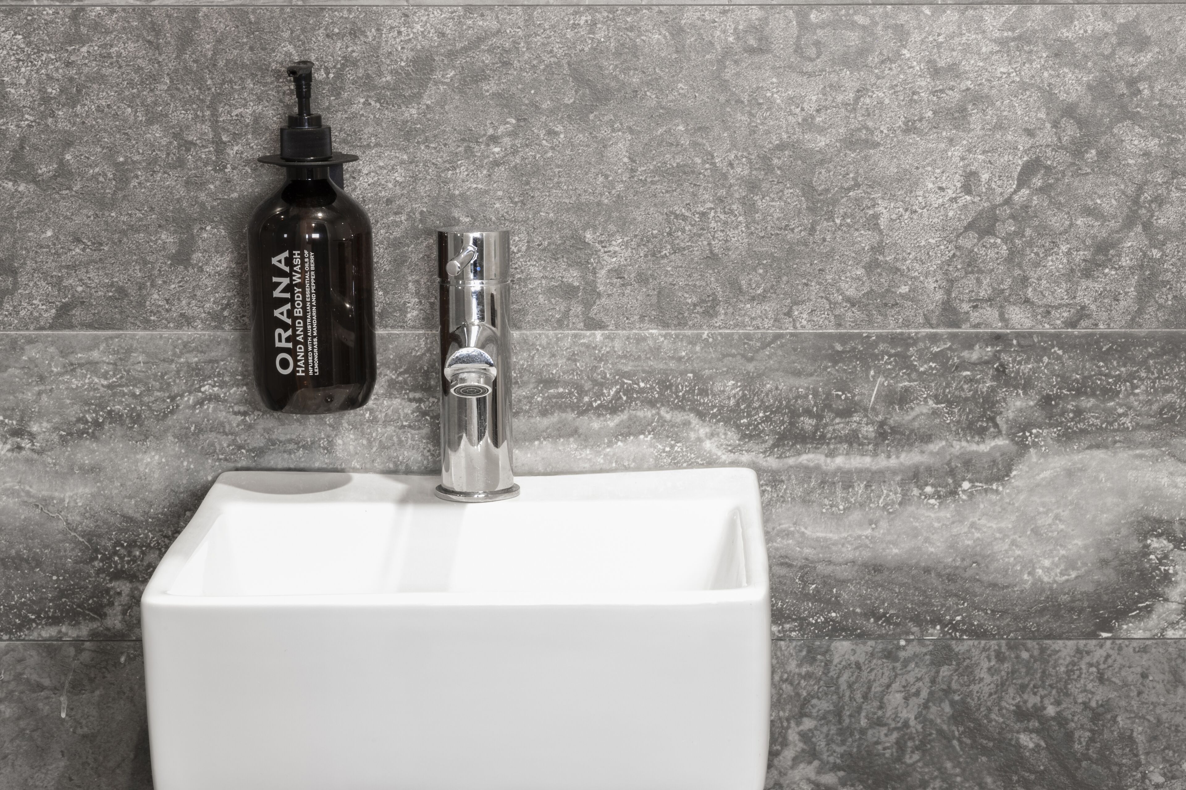 Casa de banho com lavatório branco, frasco de gel de banho e corpo Orana e torneira moderna.