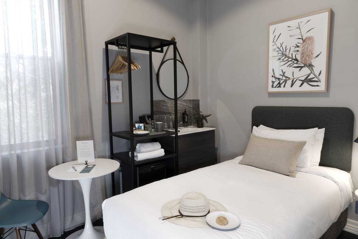 A neatly arranged hotel room with a bed, a small round table, a chair, a wardrobe, and a framed artwork on the wall.
