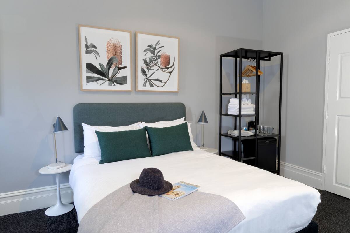 A neatly made bed with a hat and book, a nightstand with a lamp, and a black shelving unit with towels.