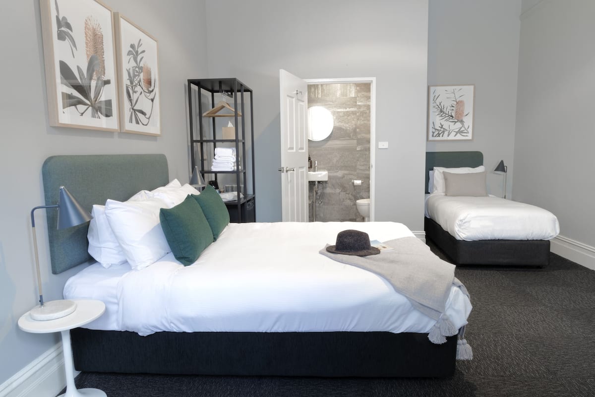 A modern hotel room with a large bed, a smaller bed, a bedside table, a wardrobe, and a bathroom visible through an open door.