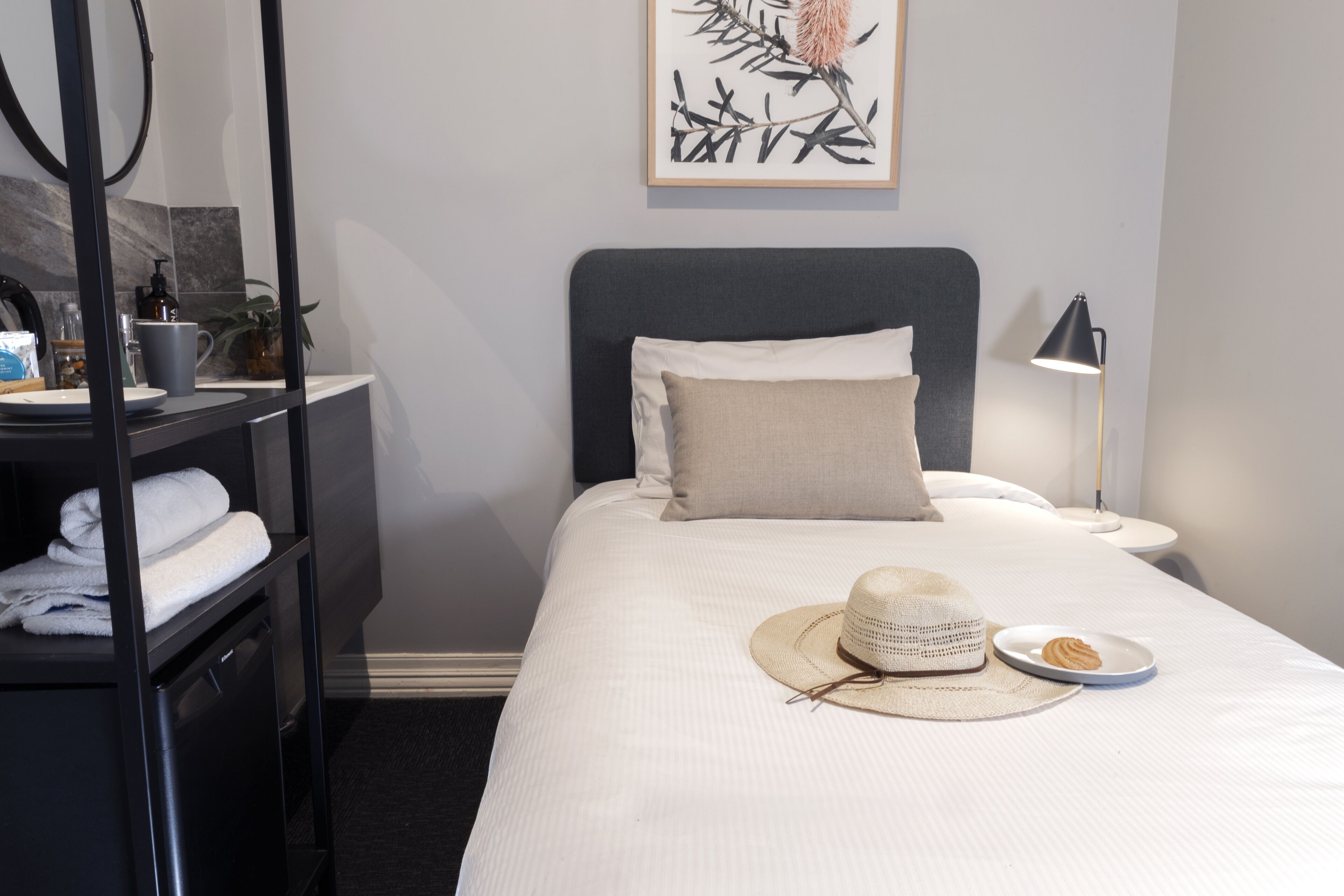 A neatly made bed with a white comforter, a beige pillow, and a straw hat on the bed. There is a small plate with a dessert on the bed. A lamp is on the nightstand beside the bed.