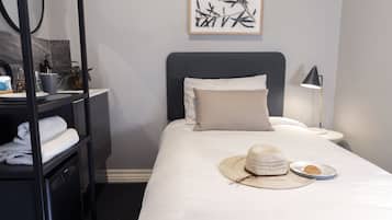 A neatly made bed with a white comforter, a beige pillow, and a straw hat on the bed. There is a small plate with a dessert on the bed. A lamp is on the nightstand beside the bed.
