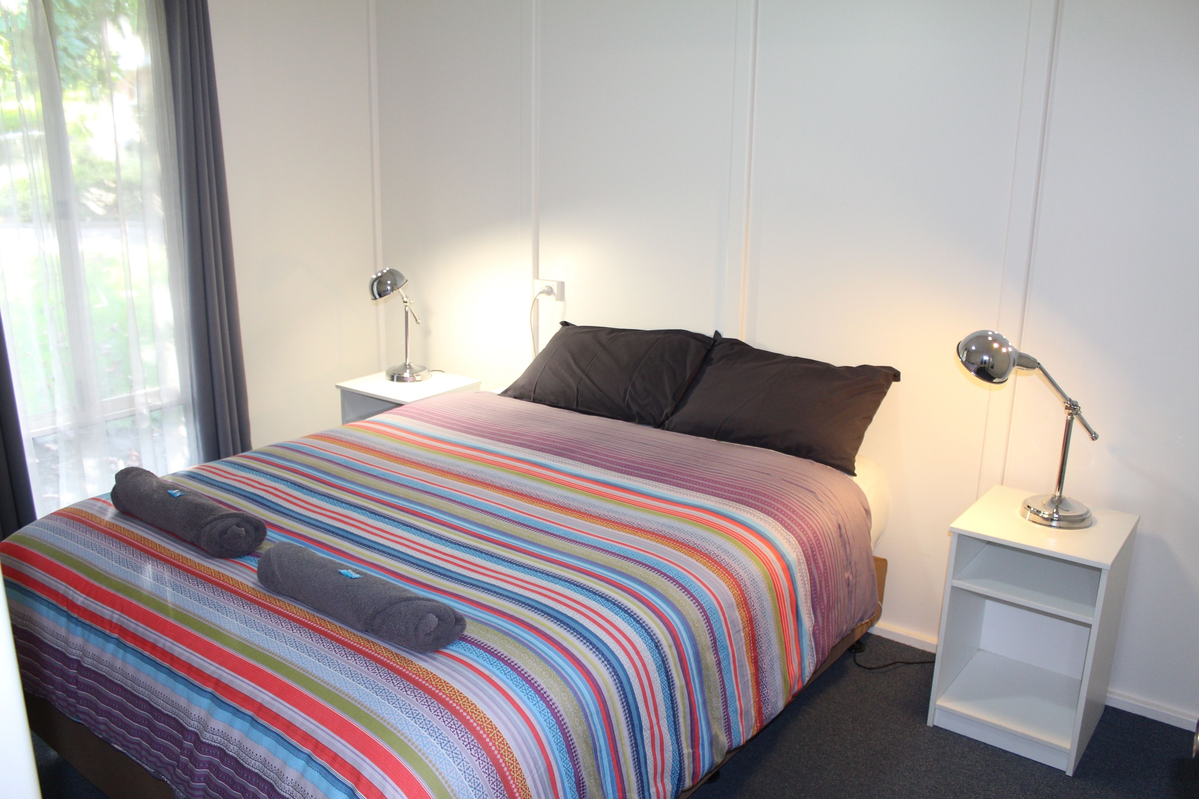 Cama bem arrumada com colcha listrada e colorida e dois travesseiros, mesa de cabeceira com abajur e janela com cortinas.