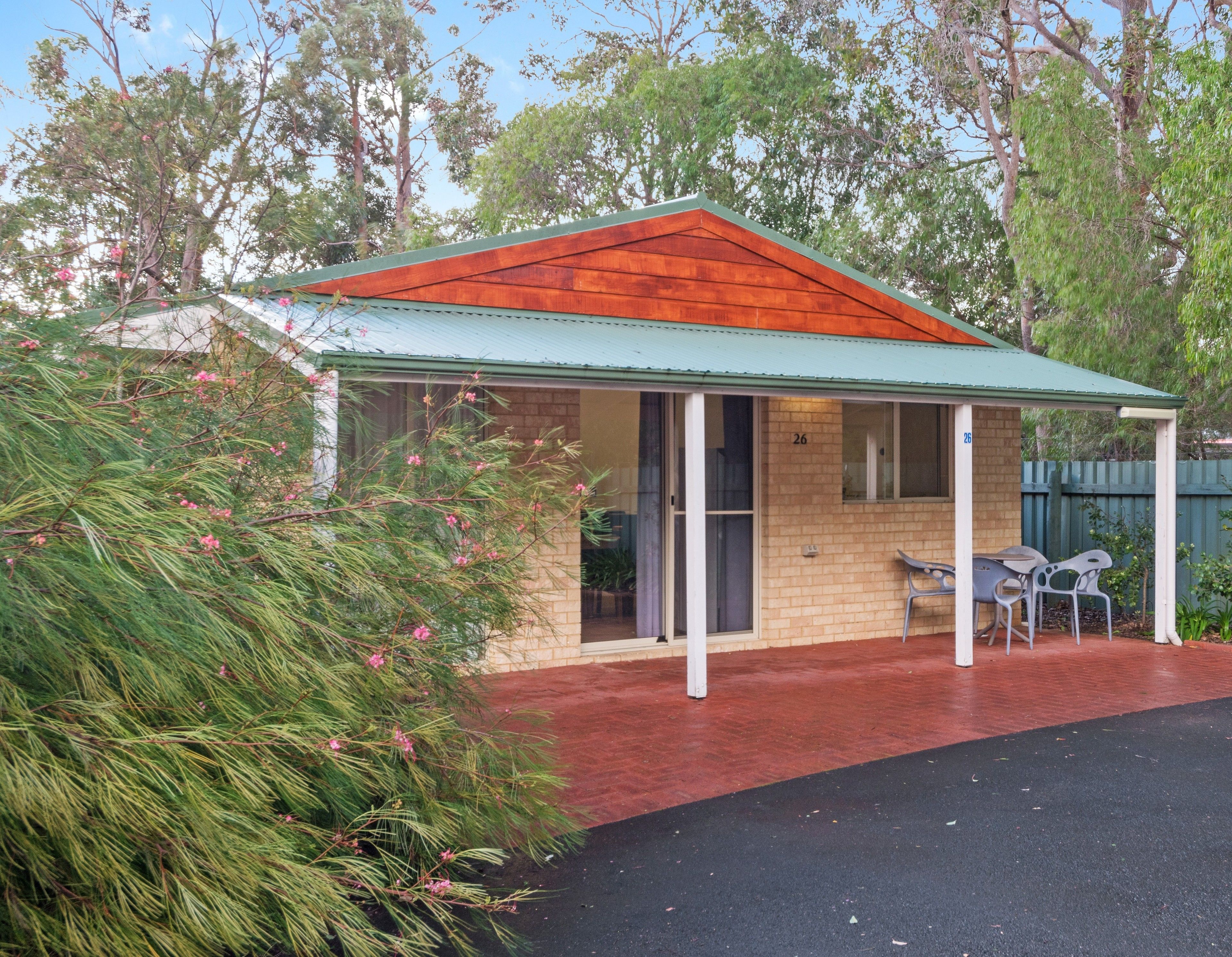 A small, single-story building with a green roof and a brick exterior. It features a covered porch with two chairs and a table.