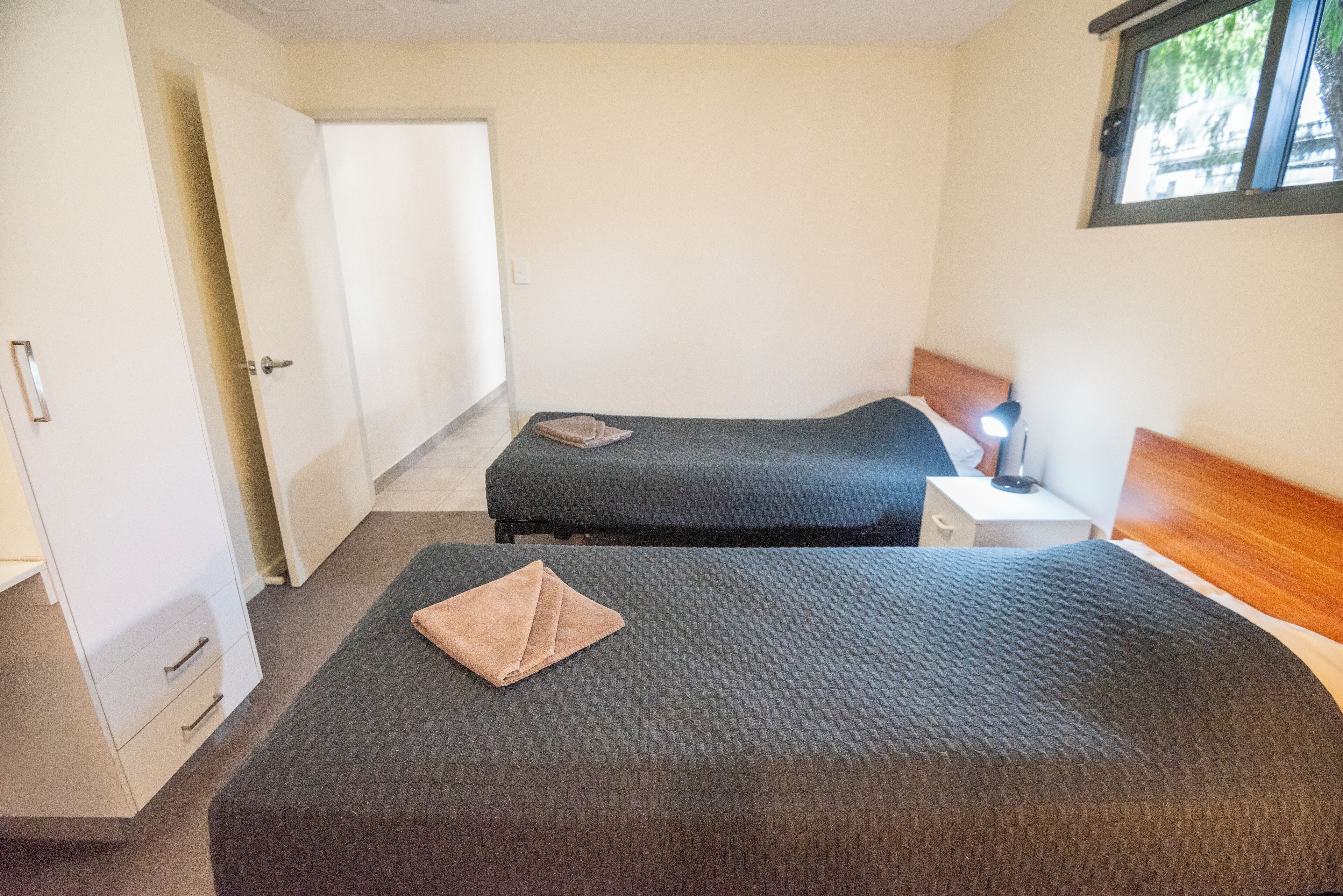Quarto simples e limpo com duas camas, uma mesa de cabeceira, um abajur e uma janela.