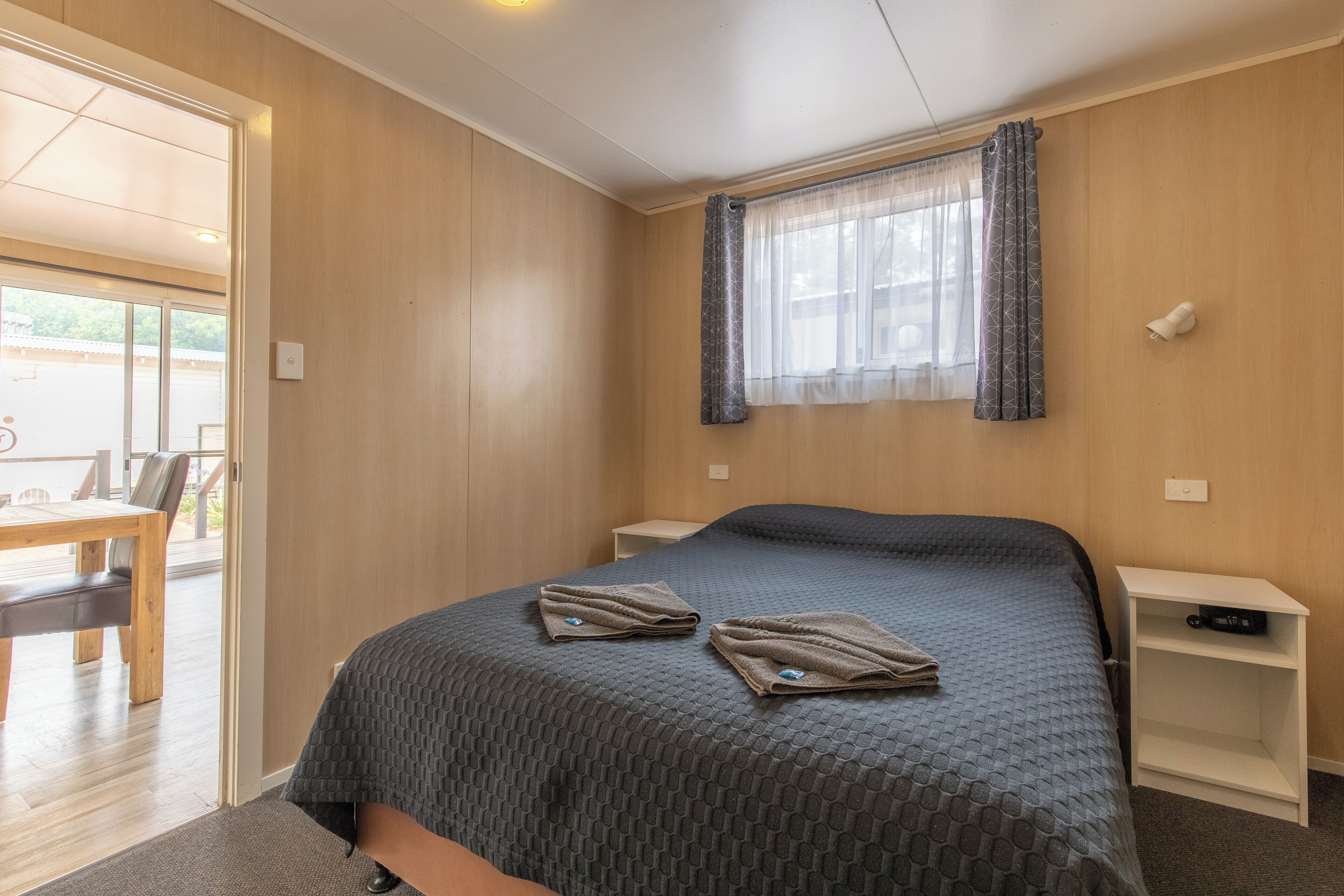 A bedroom with a bed, a window with curtains, a bedside table, and a chair visible through an open door.