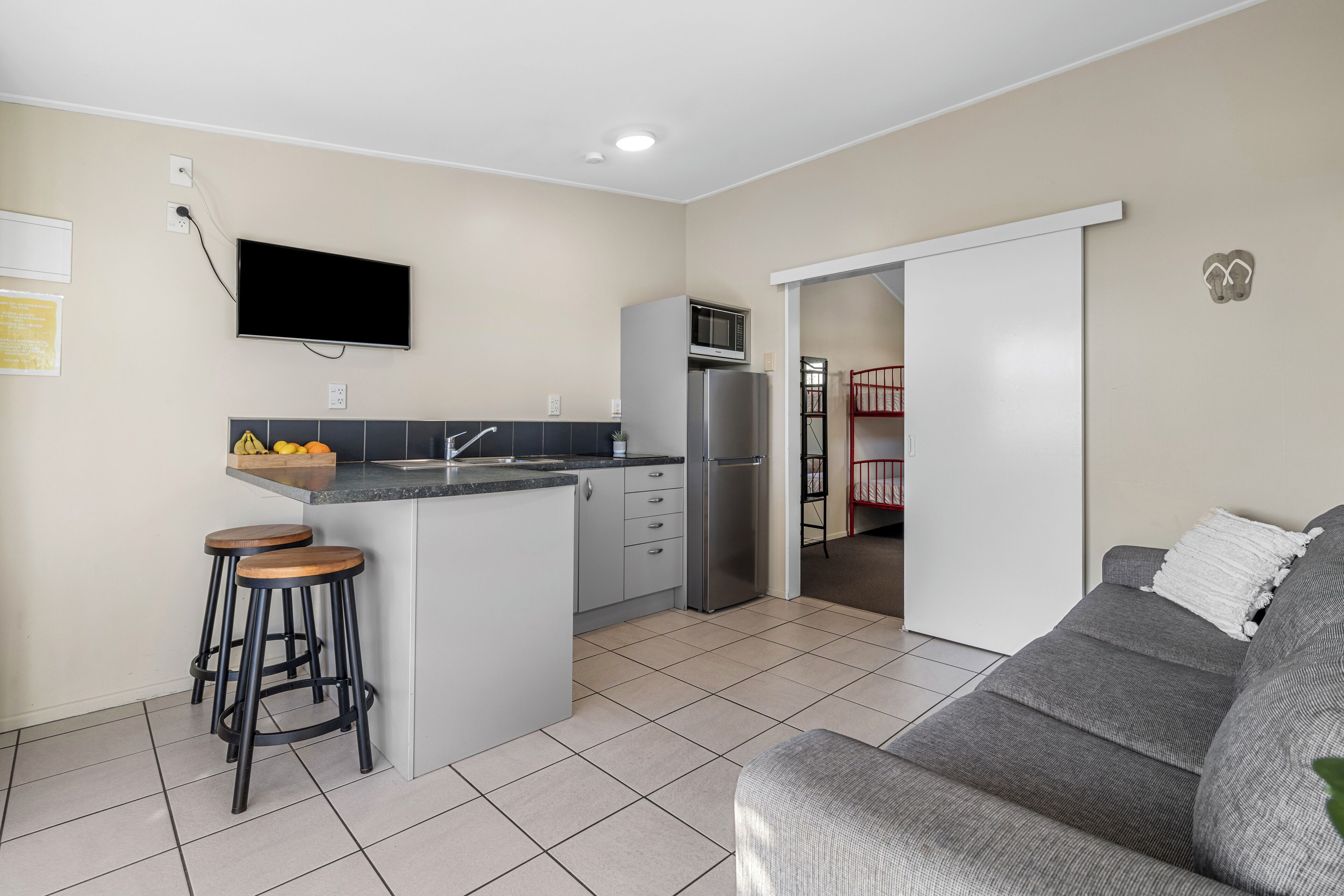 Un espacio habitable compacto con kitchenette, un sofá gris y un televisor montado en la pared.