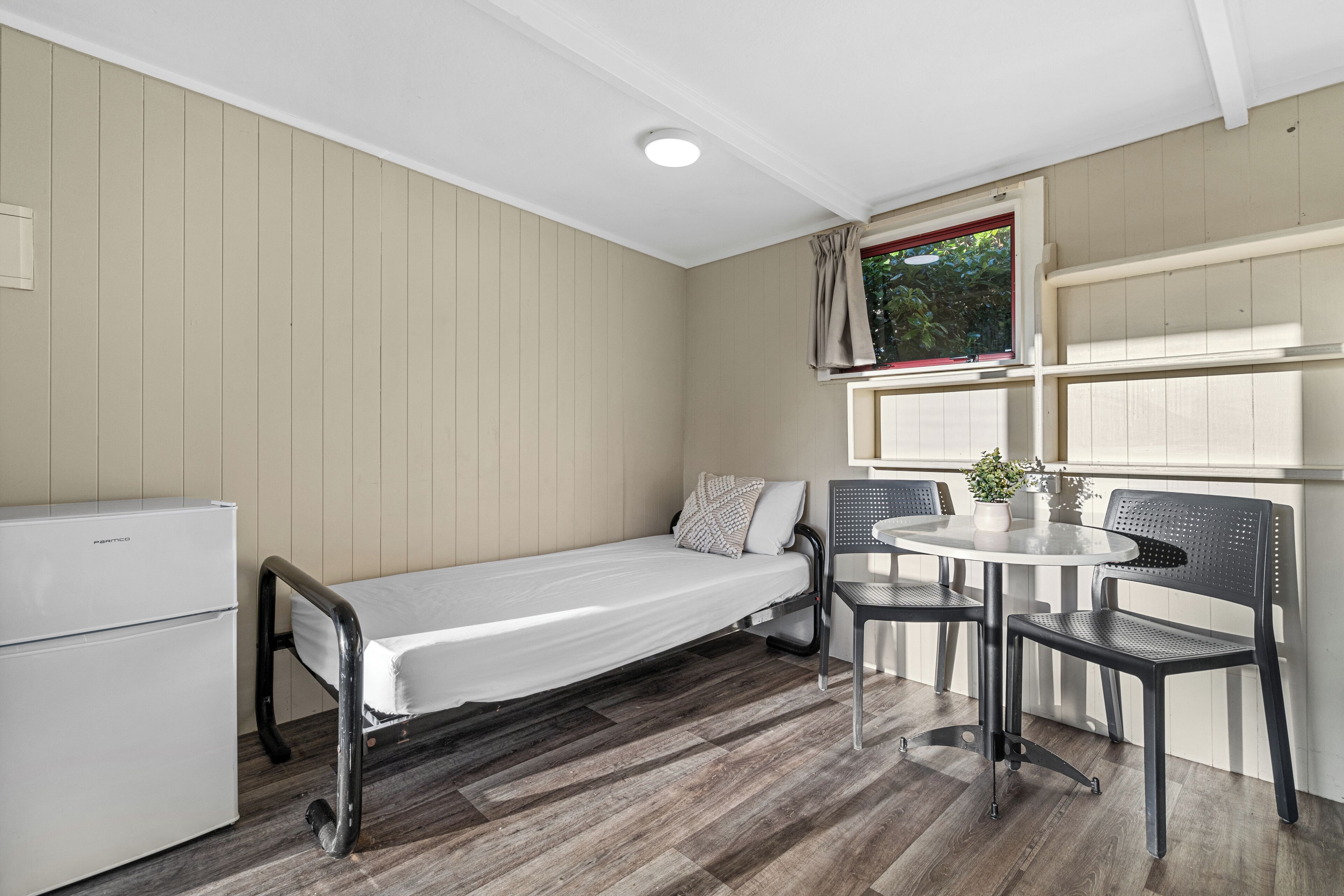 Une pièce de taille modeste comprenant un lit simple, une petite kitchenette, une table à manger avec des chaises et une fenêtre avec des rideaux.