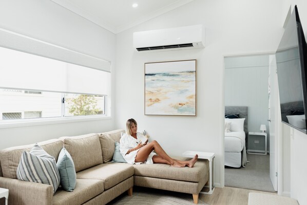 A modern, well-lit room with a large window, a beige sectional sofa, a bedside table, a bed, and a mounted artwork.