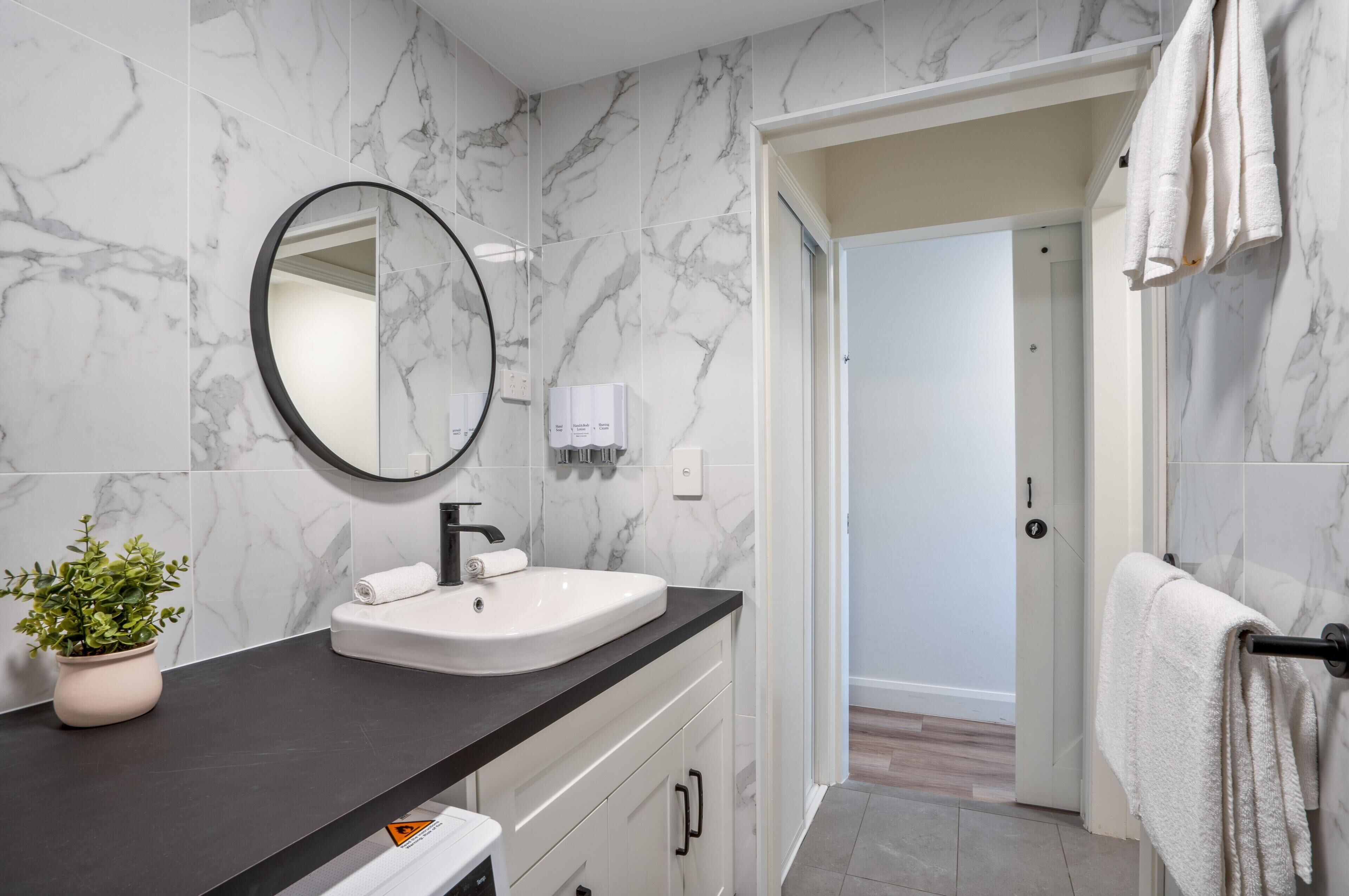 A bathroom with a marble wall, a round mirror, a sink, a towel rack, and a potted plant.