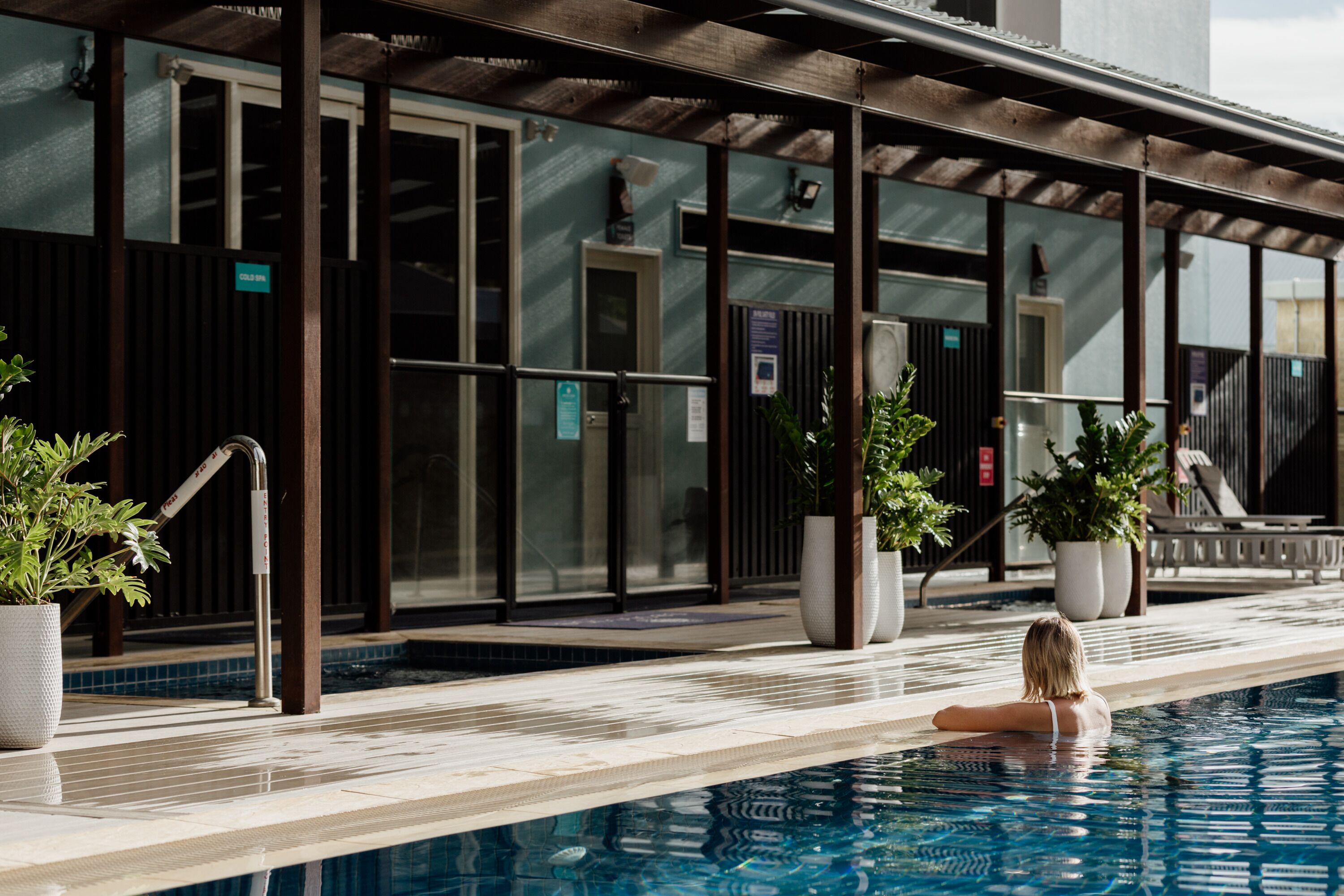 A swimming pool with a tiled deck, potted plants, and a covered area with glass doors.